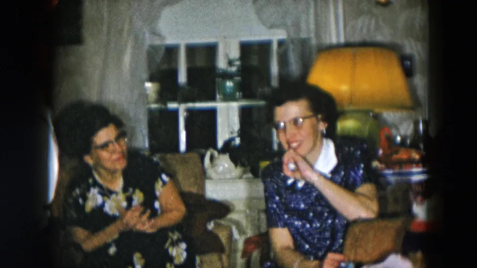 Two women in conversation in a 1950s domestic living room; woman in dark floral dress gesturing at right, older woman with lighter hair at left, amber table lamp visible behind them.