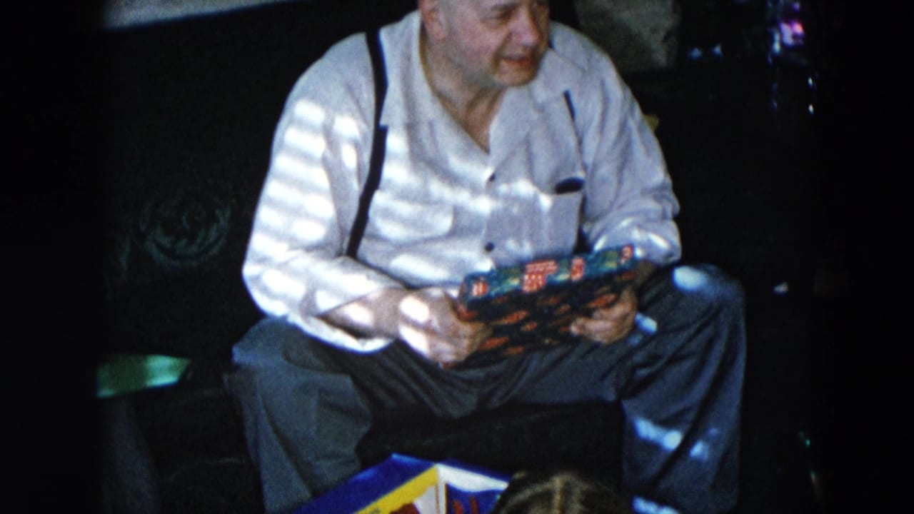 Older man in white shirt and dark suspenders examines a colorful wrapped gift in a dimly lit living room interior, Hicksville, New York, circa 1956, 8mm home movie.