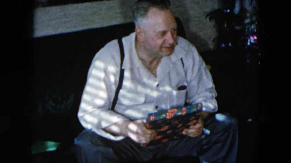 Older man in white shirt and dark suspenders holds a colorful wrapped package in a domestic interior; possible evergreen visible at upper right edge, Hicksville, New York, circa 1956, 8mm.