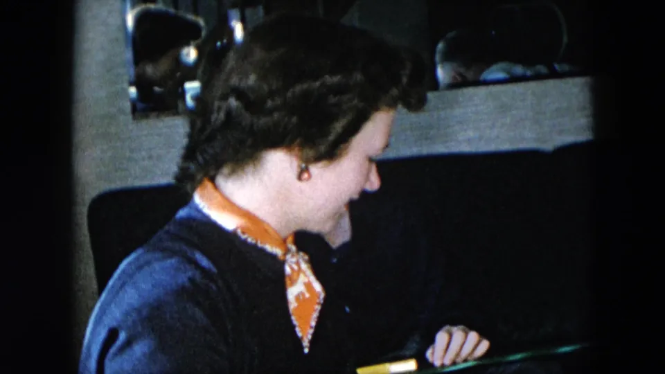 Woman wearing dark navy sweater with orange-patterned scarf and drop earring, seated in profile in a domestic interior, Hicksville, New York, circa 1956, 8mm home movie.