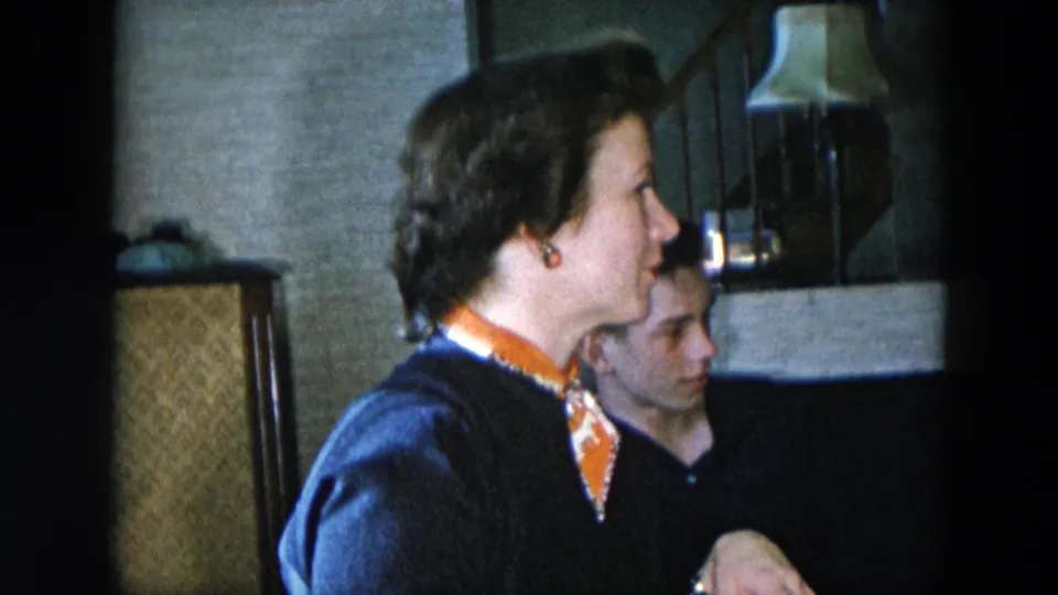 Woman in dark navy sweater and orange-patterned scarf seated in profile in a mid-century living room, Hicksville, New York, circa 1956, 8mm home movie.