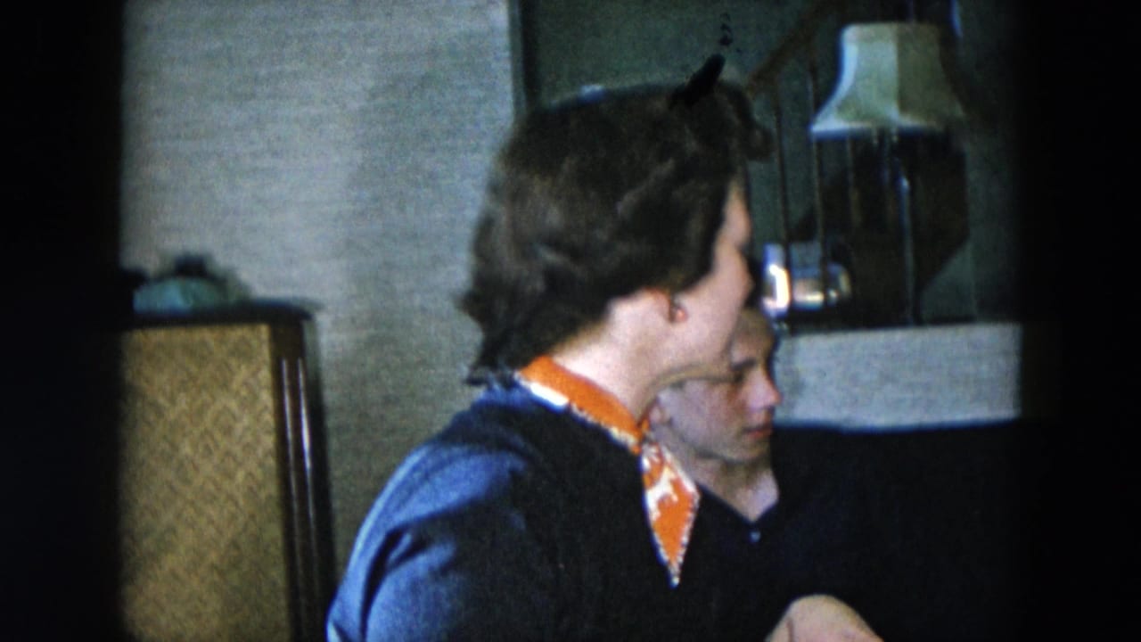 Woman in navy cardigan and orange printed neckerchief, seated in profile in what appears to be a 1956 Hicksville, New York home interior; man partially visible beside her.