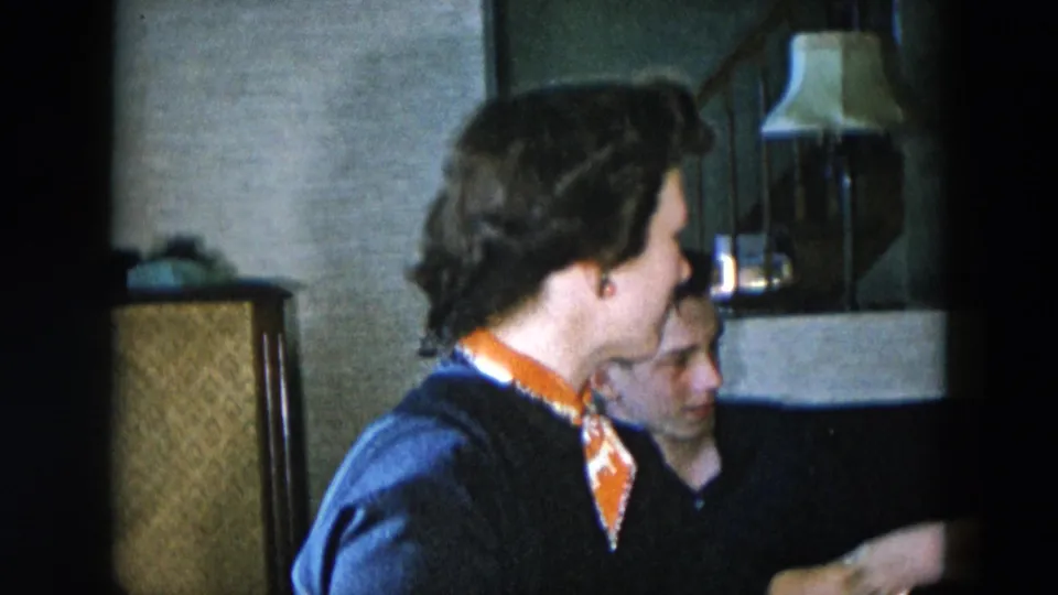 Close medium shot of woman in navy cardigan and orange neckerchief in profile, man partially visible beside her, in a mid-century domestic interior consistent with 1950s decor.