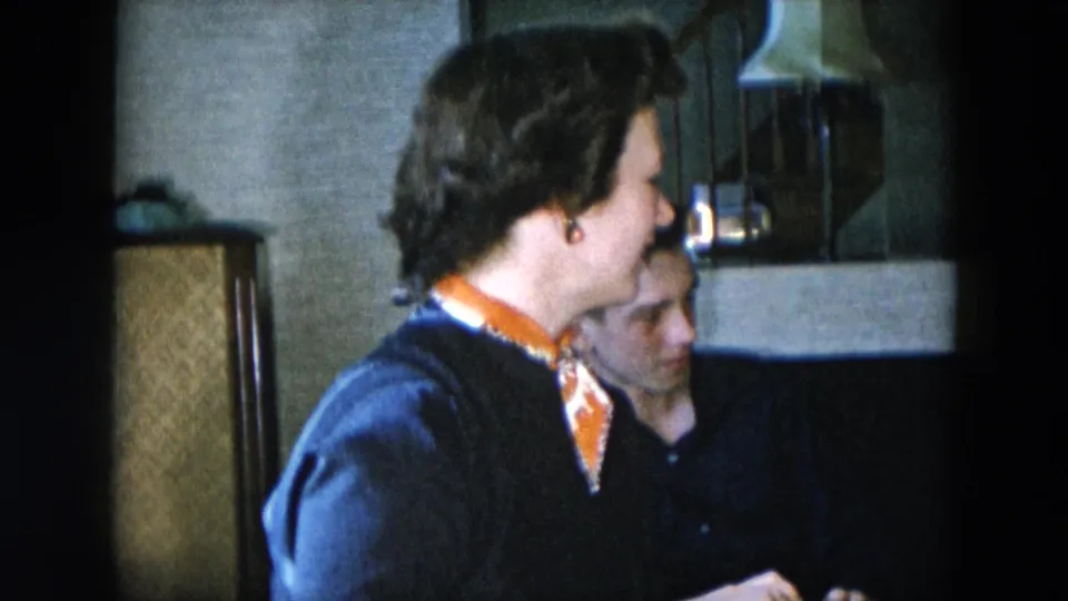 Woman animated mid-conversation in dark cardigan and orange patterned scarf, with man beside her in a 1950s domestic interior setting.