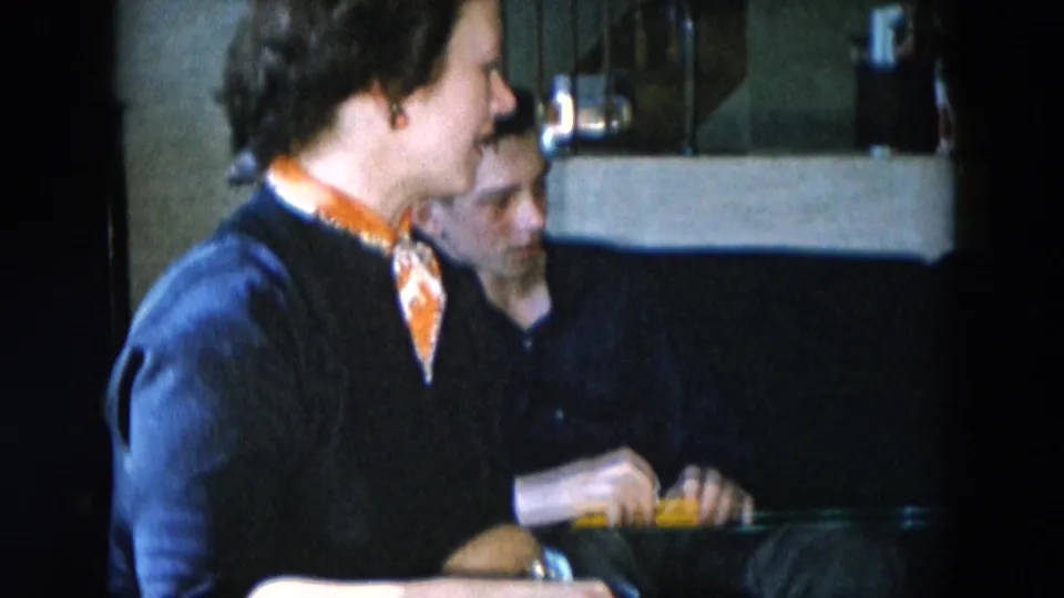 Woman and man in close interior shot in what appears to be a 1950s residential setting; man is partially visible and appears to be handling a small object.
