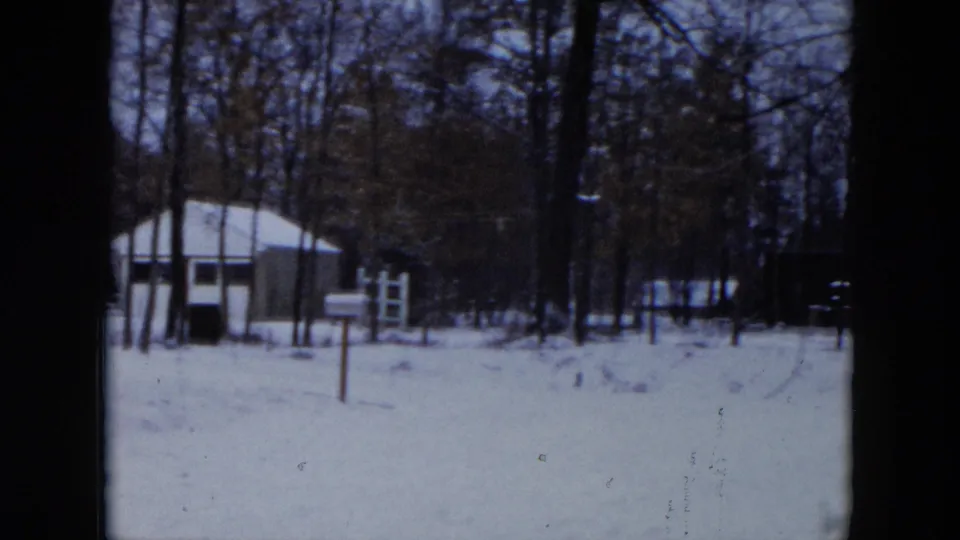 Wide view of snow-blanketed suburban yards in Hicksville, New York, circa 1956 — bare trees, distant white house, and flat winter light captured on 8mm film.