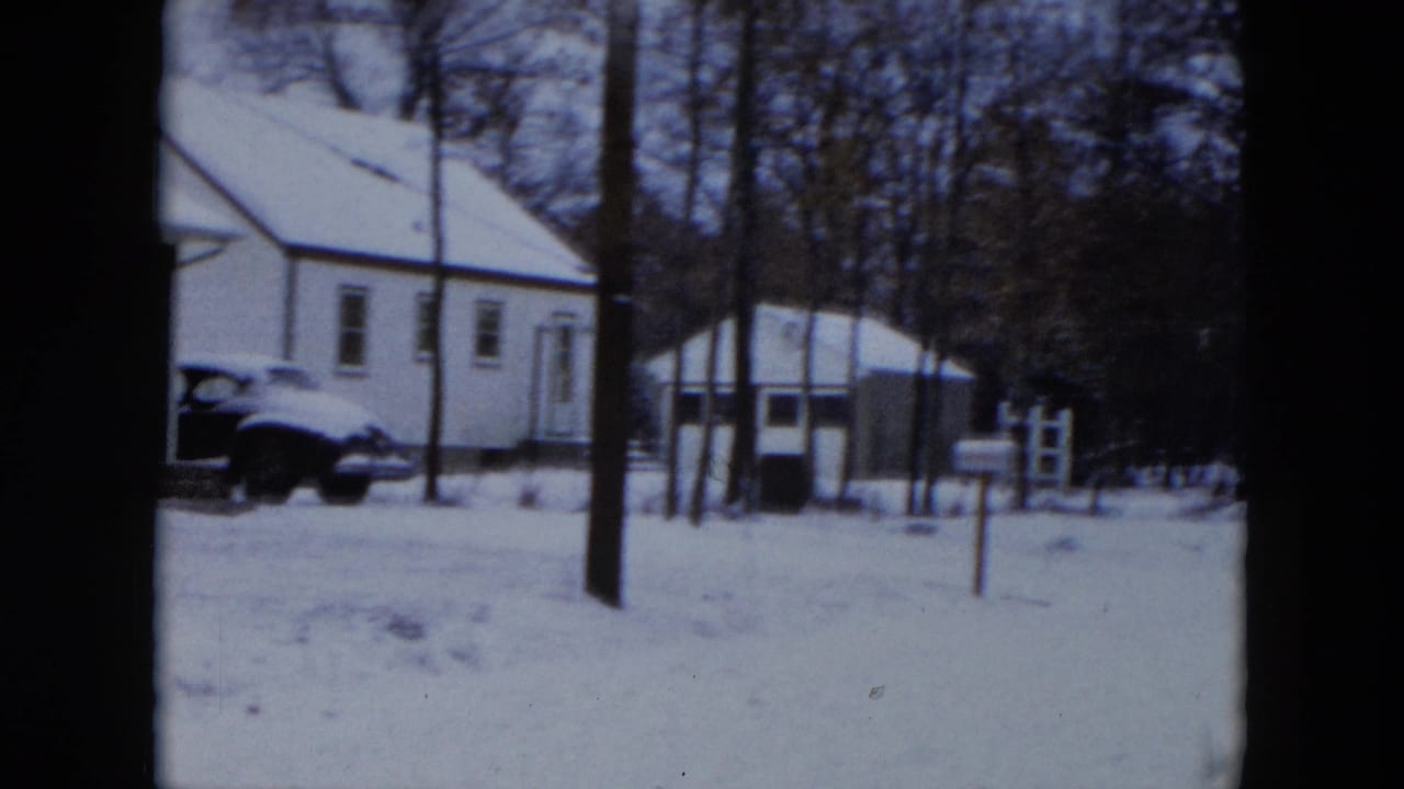 White house with snow-covered roof and a dark-colored 1950s sedan parked in the driveway, Hicksville, New York, circa 1956, on 8mm film.