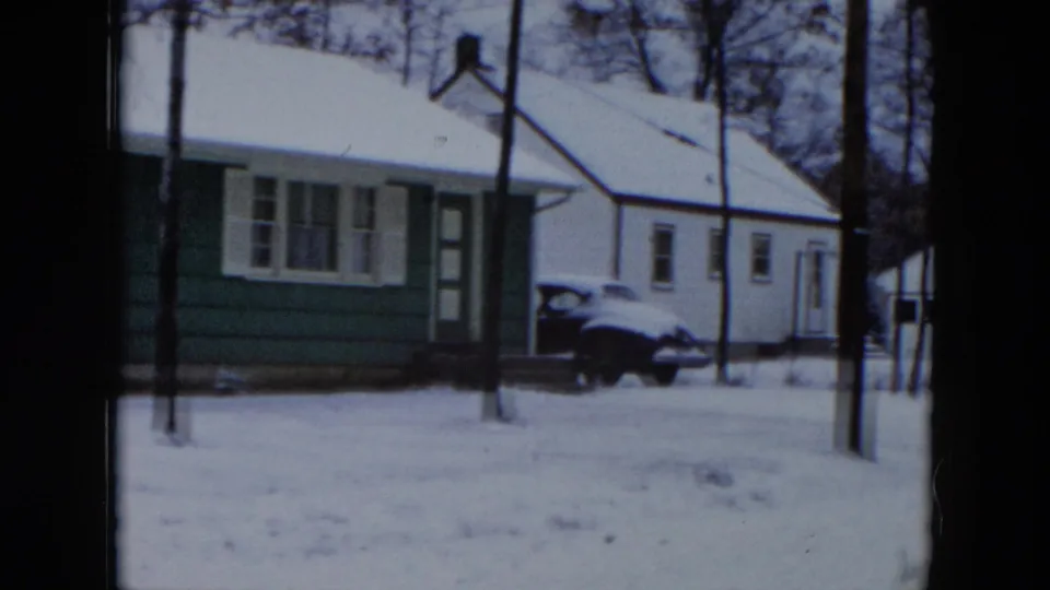 Green ranch-style house with white trim and snow-covered roof, with a dark 1950s car parked at left, in a winter scene in Hicksville, New York, circa 1956.