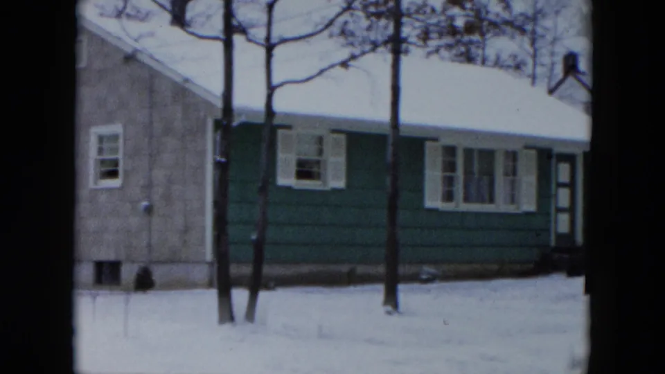 Green house and adjacent gray structure with snow-covered roofs and bare winter trees, Hicksville, New York, circa 1956, on 8mm film.