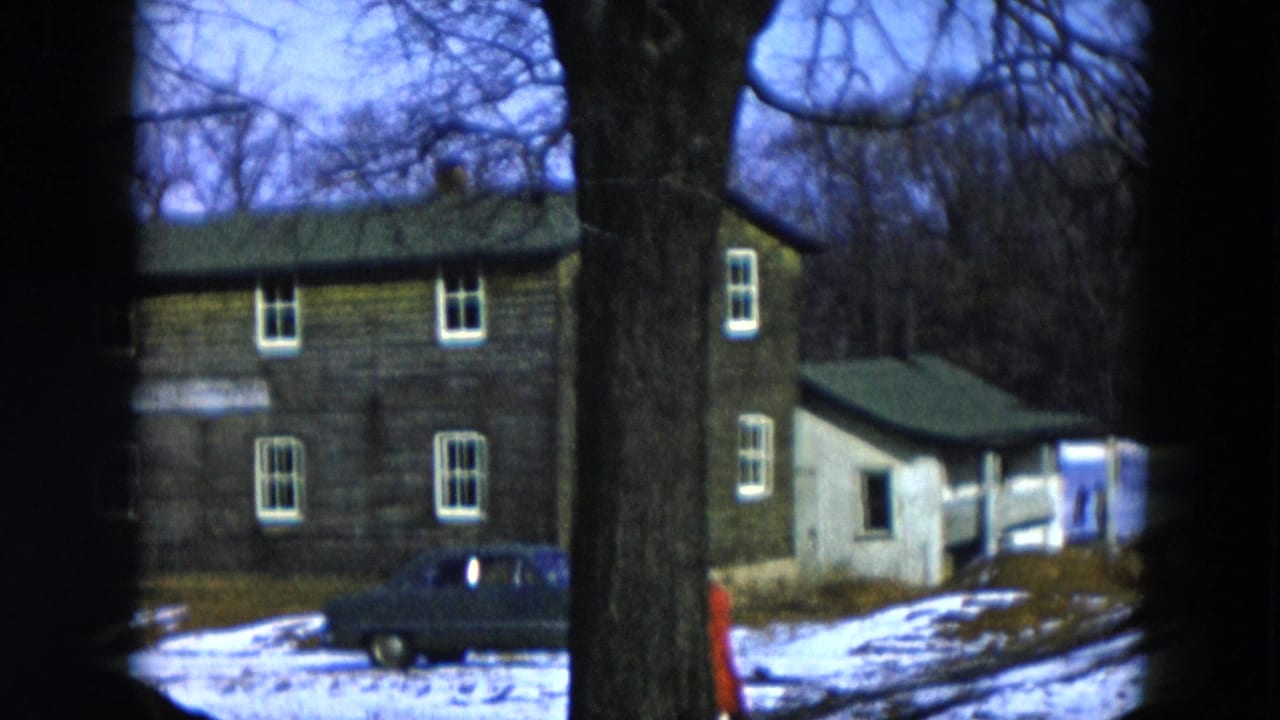A weathered two-story house in winter with a large tree trunk in the foreground and a dark sedan parked nearby; a figure in a red coat is visible at the lower right edge of frame.