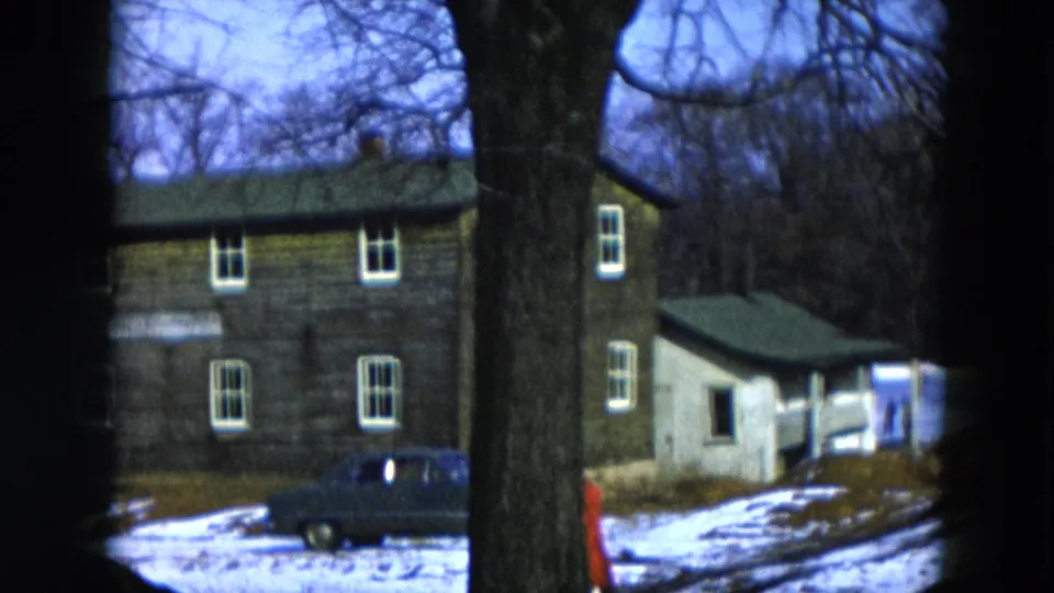 8mm home movie frame, 1956 Hicksville New York: two-story clapboard house in winter, vintage dark sedan parked at left, person in red coat at lower right, large foreground tree.