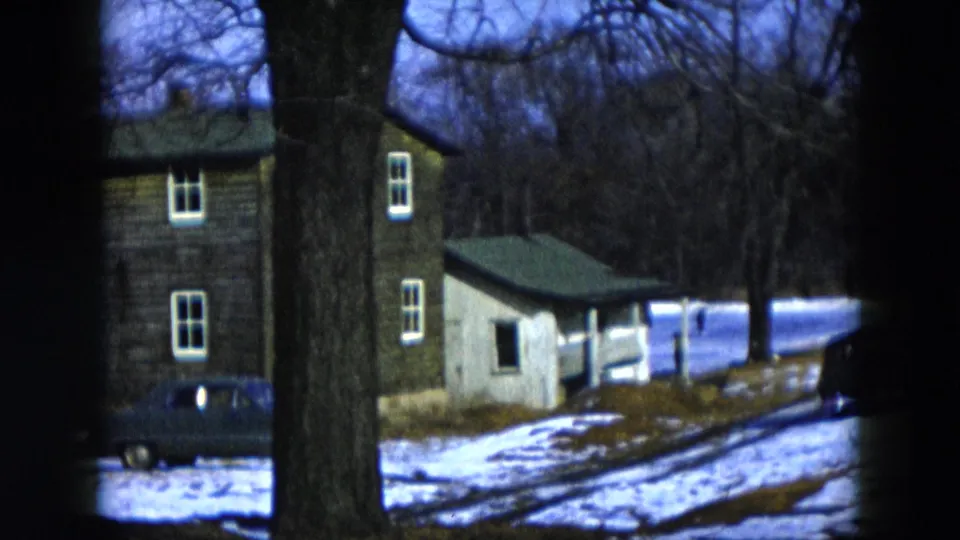 8mm pan frame, 1956 Hicksville New York: two-story house and small outbuilding with green roof behind foreground tree, dark sedan on patchy snow, bare trees under blue-toned sky.