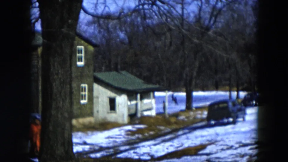 Mid-pan 8mm frame, 1956 Hicksville New York: outbuilding prominent at center, red-coated figure at left frame edge, dark vehicle on snow, bare winter trees filling the background.