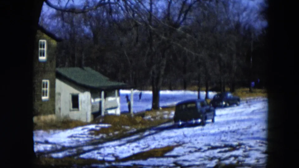 End-of-pan 8mm frame, 1956 Hicksville New York: snowy residential yard with outbuilding, dark sedan at right, and dense bare winter trees against a blue-toned overcast sky.