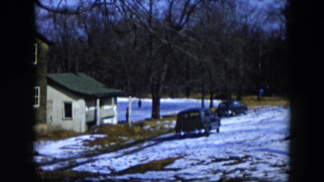Snow-covered pond area with a small outbuilding and a dark vintage car in the foreground, bare winter trees filling the background.