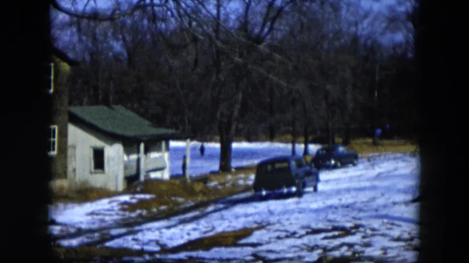 8mm home movie frame, Hicksville, New York, 1956; snow-covered skating pond with a small green-roofed outbuilding and a parked mid-1950s automobile amid bare winter trees.
