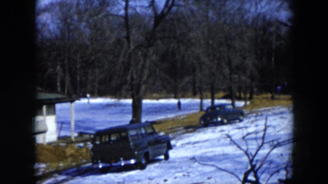 Mid-1950s station wagon parked on a snowy roadside beside a small outbuilding, with a snow-covered open area and bare trees beyond.