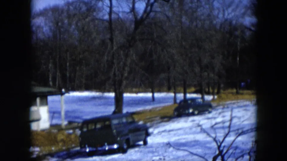 8mm home movie frame, 1956 Hicksville, New York; two mid-1950s automobiles parked beside a snow-covered pond surrounded by bare winter trees and patchy snow-and-grass ground.