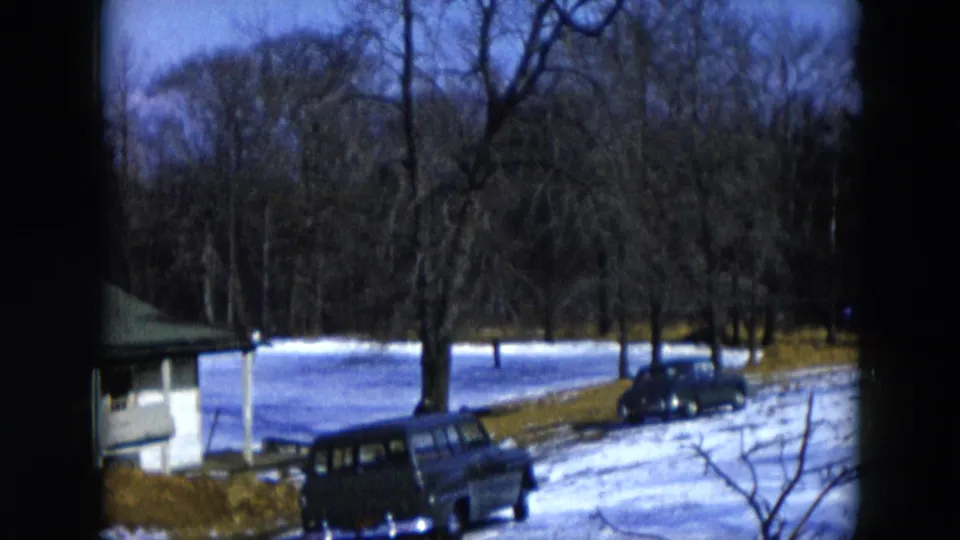 8mm home movie, 1956 Hicksville, New York; a station wagon and second vintage automobile parked beside a small green-roofed outbuilding, snow-covered pond and bare winter trees in the background.
