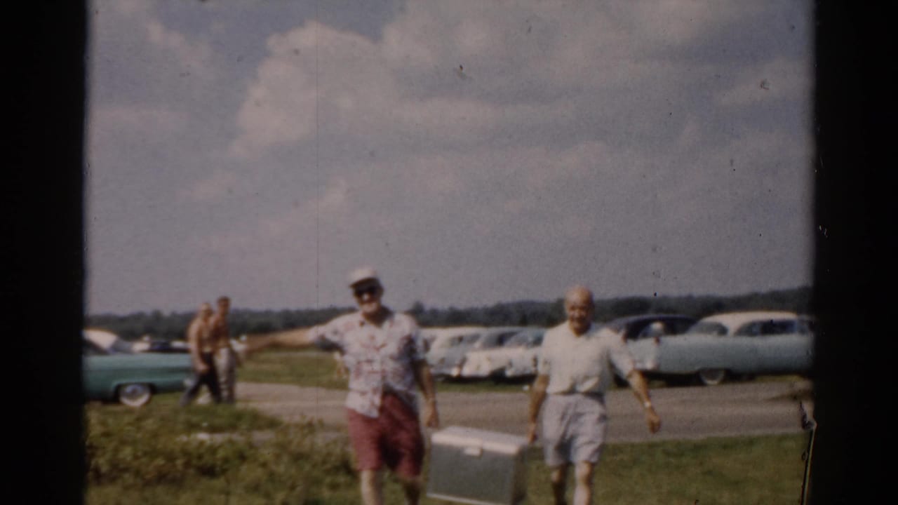 Two men in summer casual clothes walk toward camera across a grassy field; a row of 1950s American cars is parked in the background.