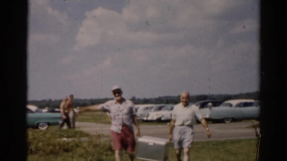 Two adult men in shorts and short-sleeved shirts approach the camera on a sunny day; a line of large American automobiles consistent with the late 1950s is parked behind them.