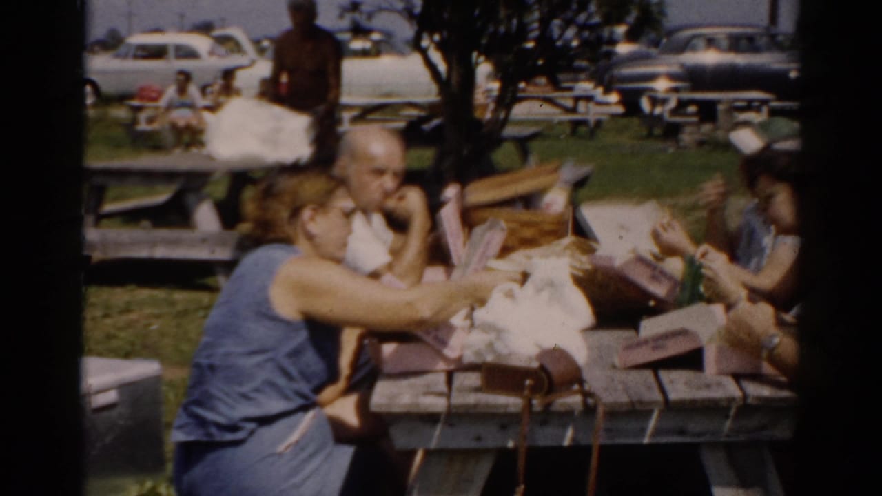 A woman in a blue top and a man interact closely at a wooden picnic table during an outdoor gathering consistent with 1950s Catskill, New York; paper containers and picnic items cover the table.