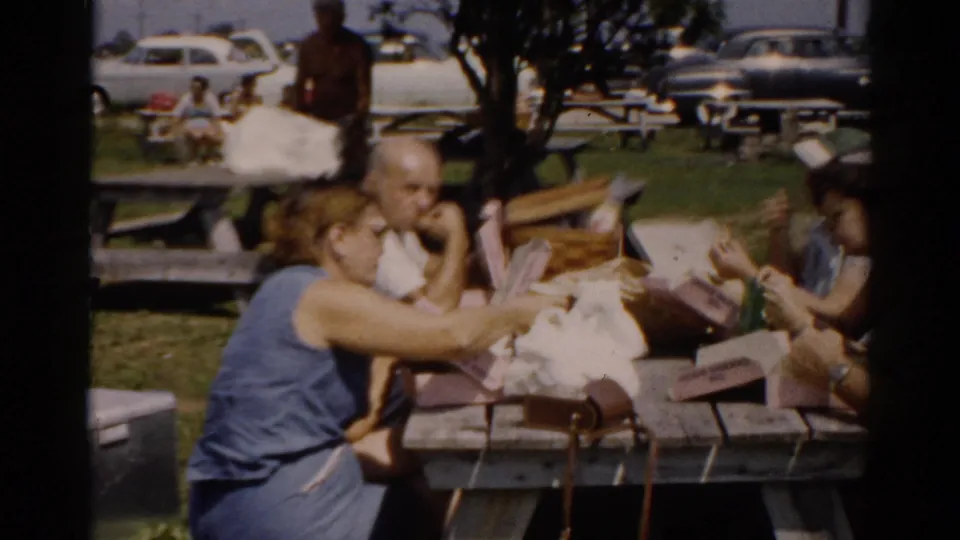 A woman in a blue top and a man interact closely at a wooden picnic table during an outdoor gathering consistent with 1950s Catskill, New York; paper containers and picnic items cover the table.