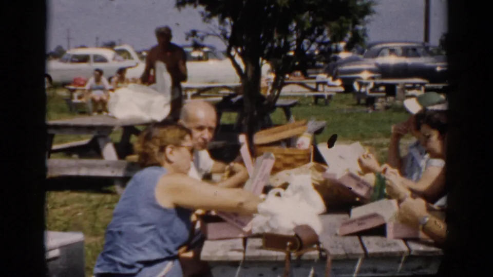 A bald man and a woman share a meal at a wooden picnic table; behind them a large open field with dozens of 1950s American automobiles and groups of people at tables stretches into the distance.