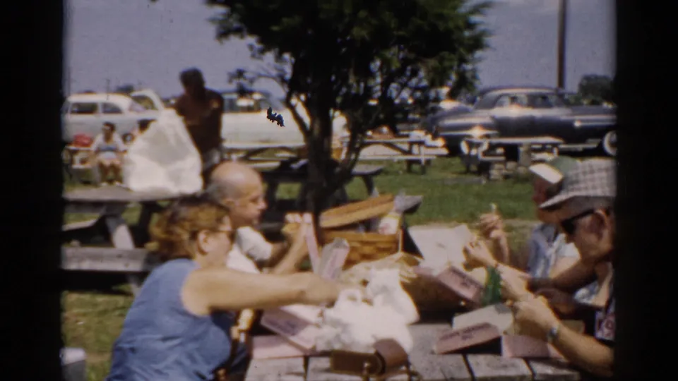 Three adults including a man in a plaid cap share a meal at a wooden picnic table at a 1950s outdoor gathering; cardboard food containers sit on the table with cars and people filling the background.