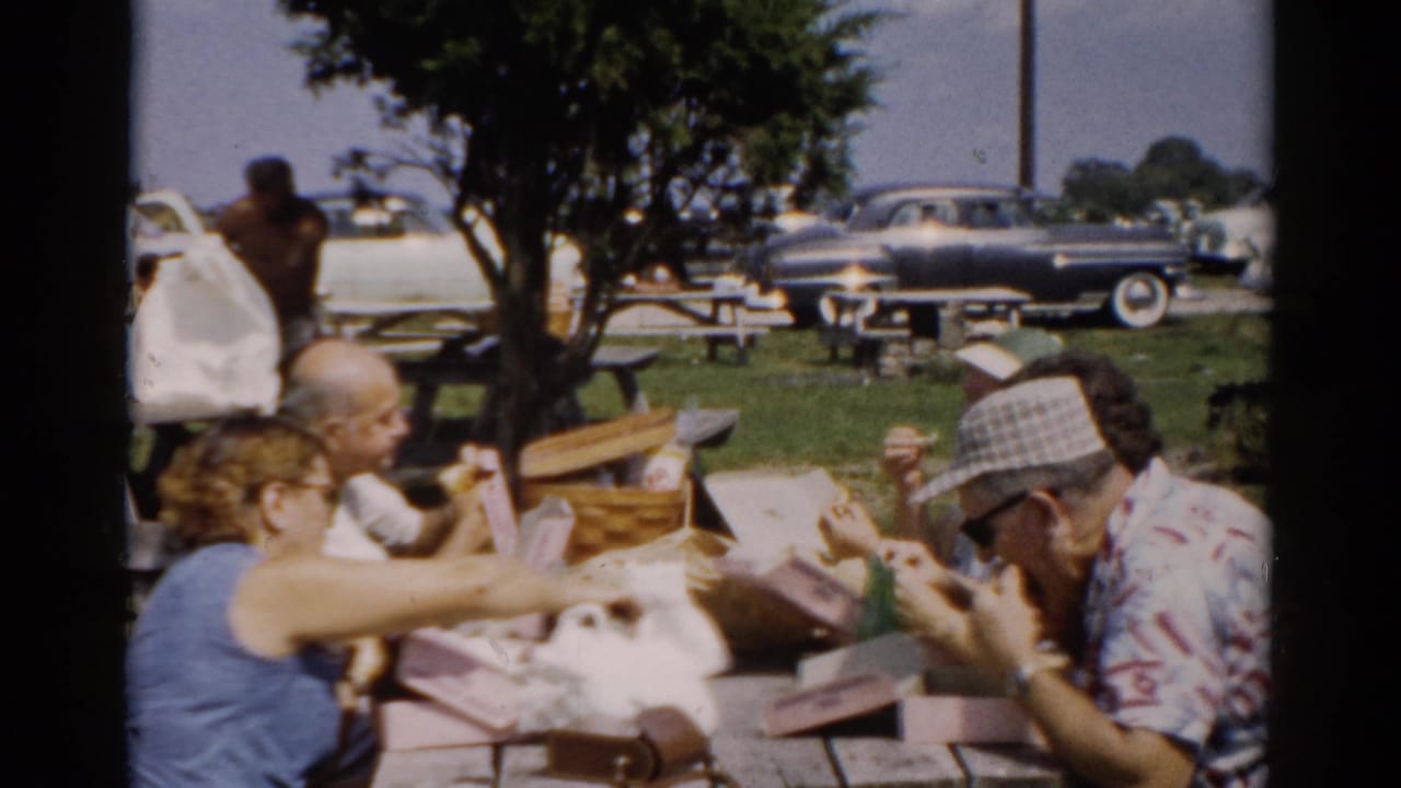 Man in plaid bucket hat and patterned sport shirt eating at a wooden picnic table, circa 1957, Catskill area, New York; late-1950s automobiles with whitewall tires visible behind.