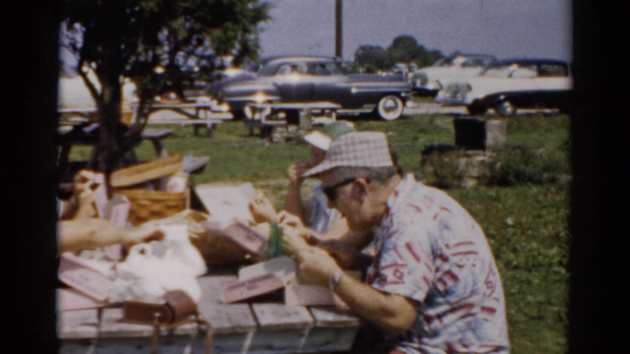 Close-up of man in plaid hat eating at a picnic table, wicker basket and wrapped food visible, vintage automobiles parked behind.