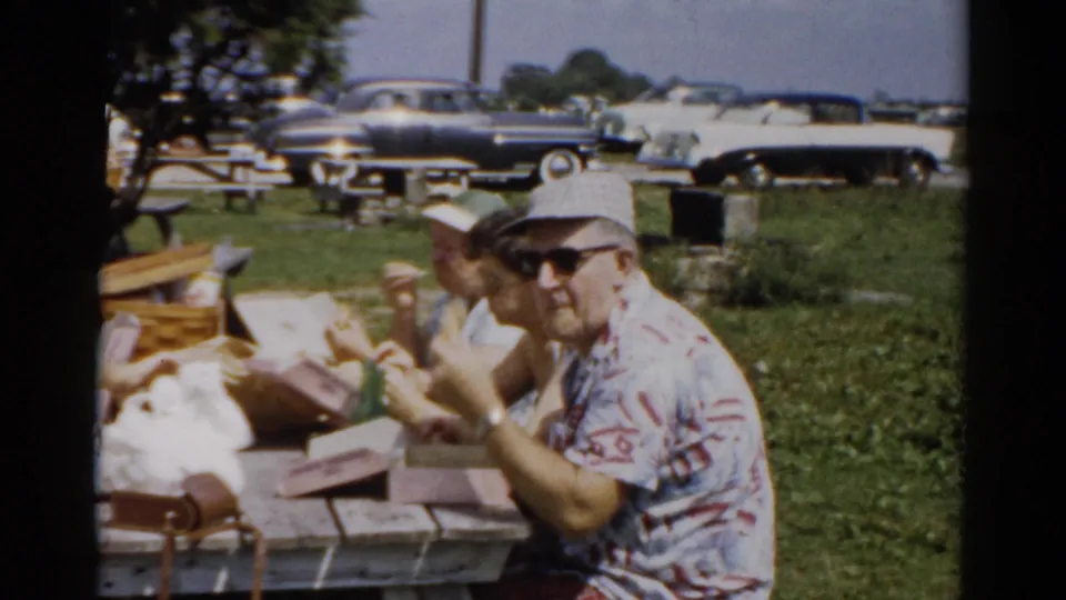 Man in plaid hat now wearing sunglasses at a summer picnic, circa 1957, Catskill area, New York; late-1950s automobiles and trees visible in background.