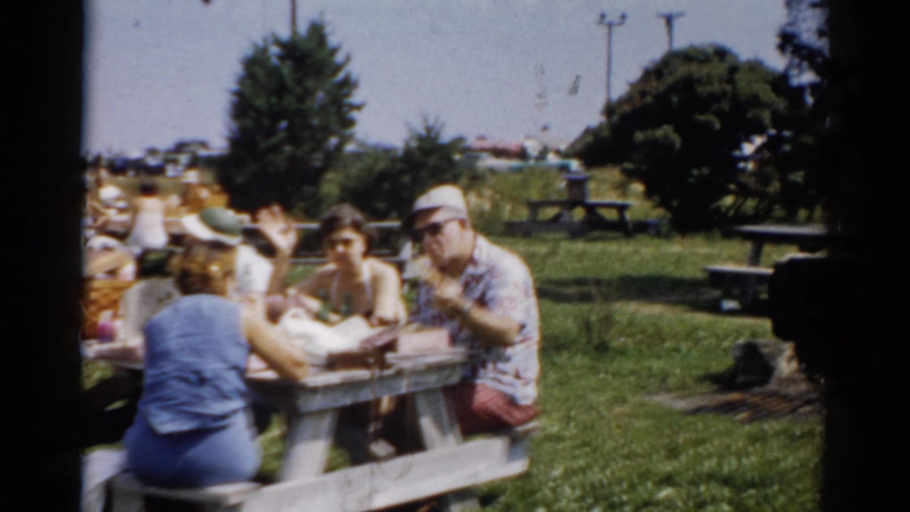 Family group eating at a public park picnic table in Catskill, New York, circa 1957; woman in blue sleeveless dress and man in patterned sport shirt and cap visible.