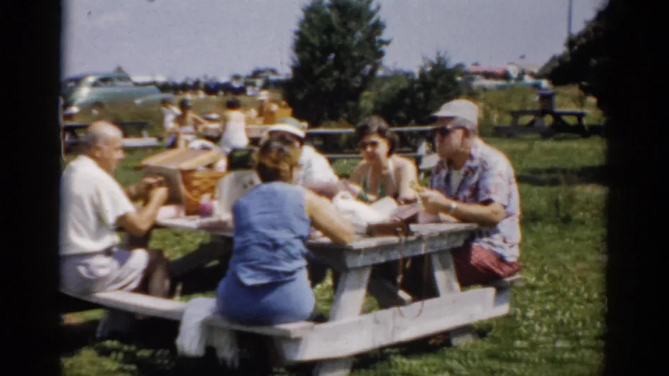 Family group at picnic table in a public park, circa 1957 Catskill, New York; wicker basket on table, multiple adults sharing food, parked vehicles faintly visible in far background.