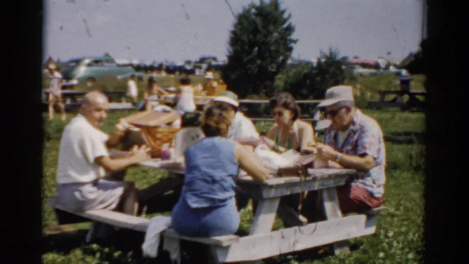 Close shot of family group at park picnic table, circa 1957; adults reaching across to share food, wicker basket visible at center, background trees and park visitors behind.