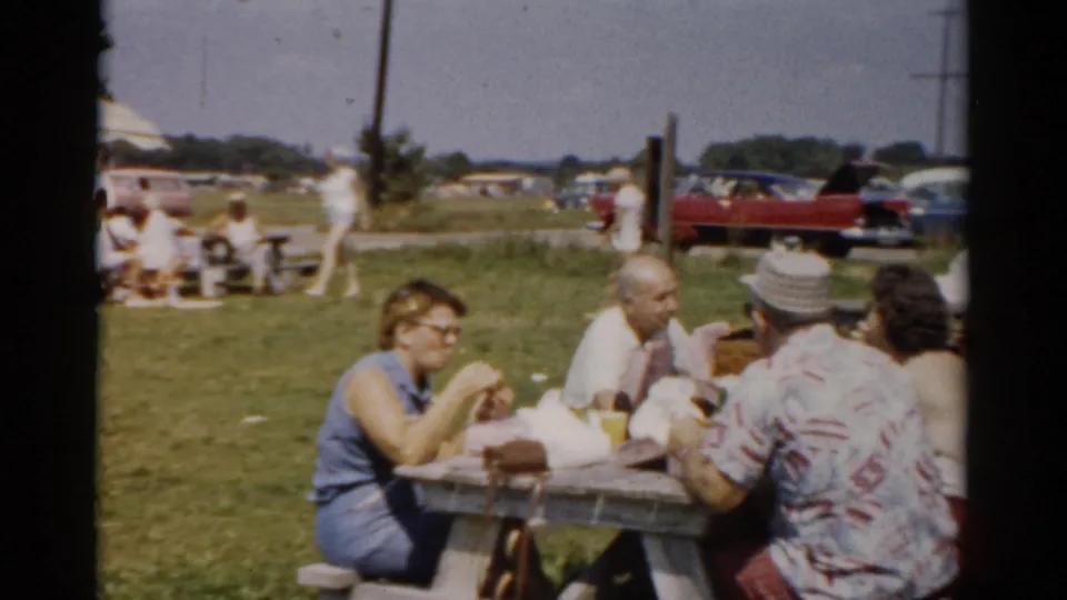 Wider shot of family group at park picnic table, circa 1957; woman with cat-eye glasses in profile, red vintage car parked in background, open grassy recreation area with additional visitors.