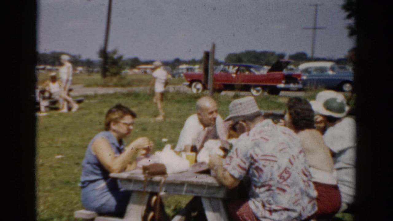 1957 Catskill, New York home-movie frame of adults eating at a picnic table on grass, with visitors and mid-century cars in the background.