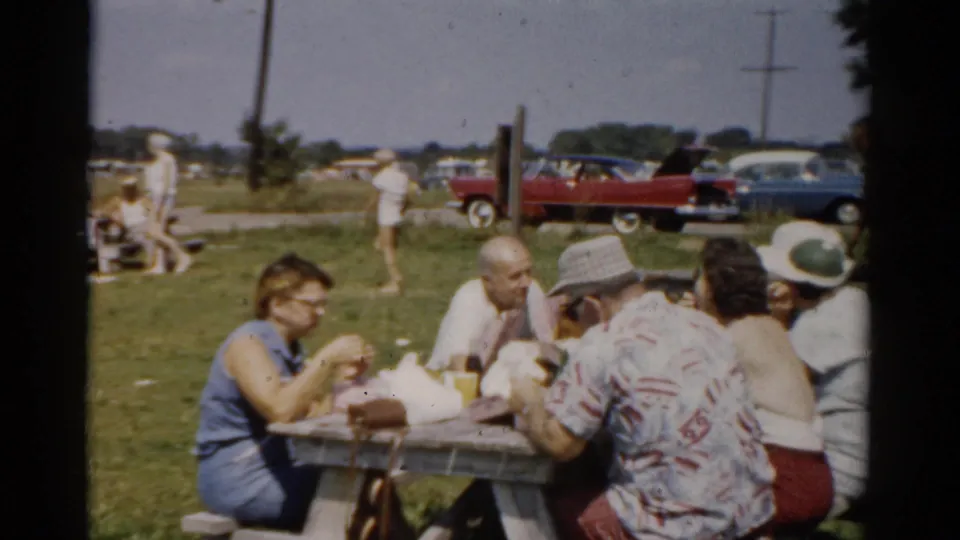 1957 Catskill, New York home-movie frame of adults eating at a picnic table on grass, with visitors and mid-century cars in the background.