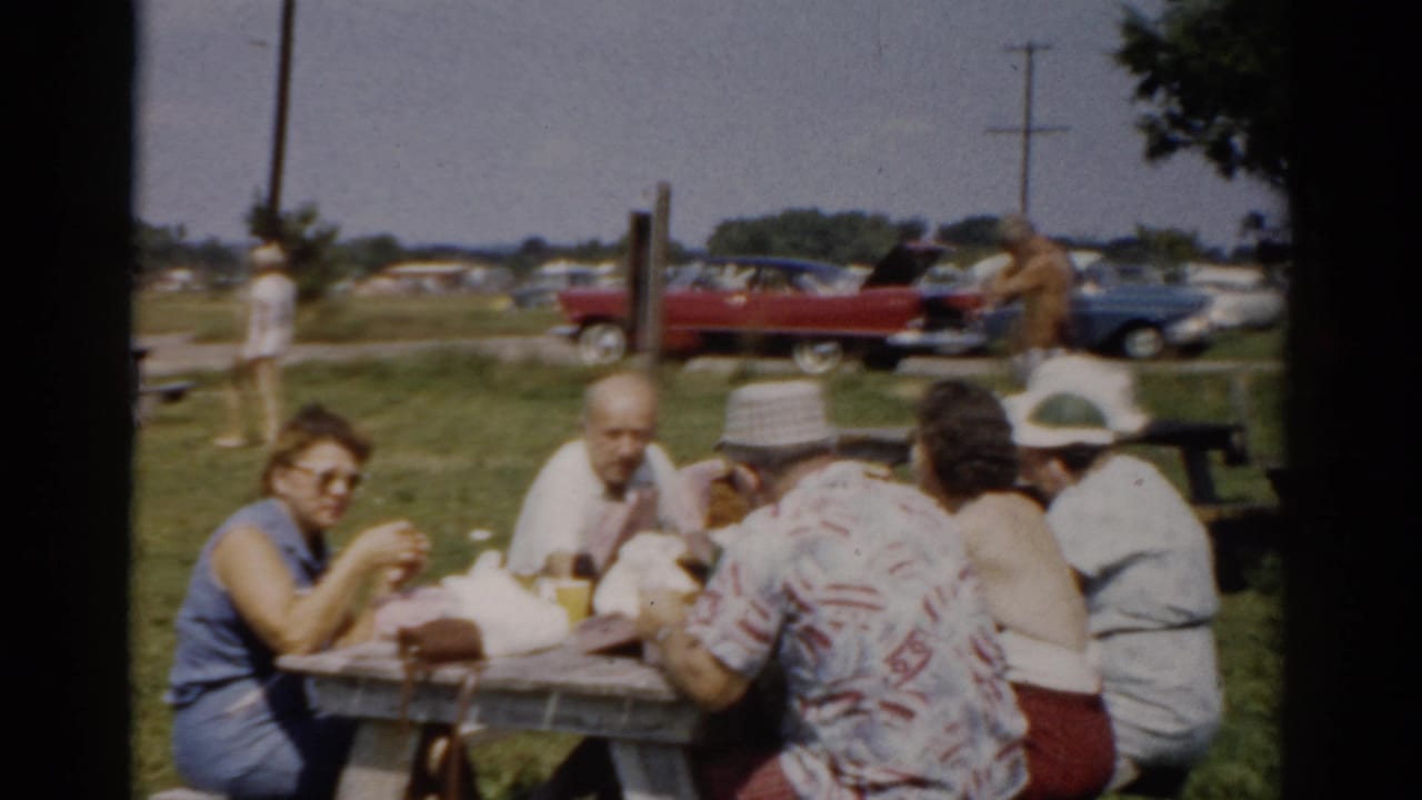 Picnic table scene with adults eating and talking on a grassy field near parked cars.