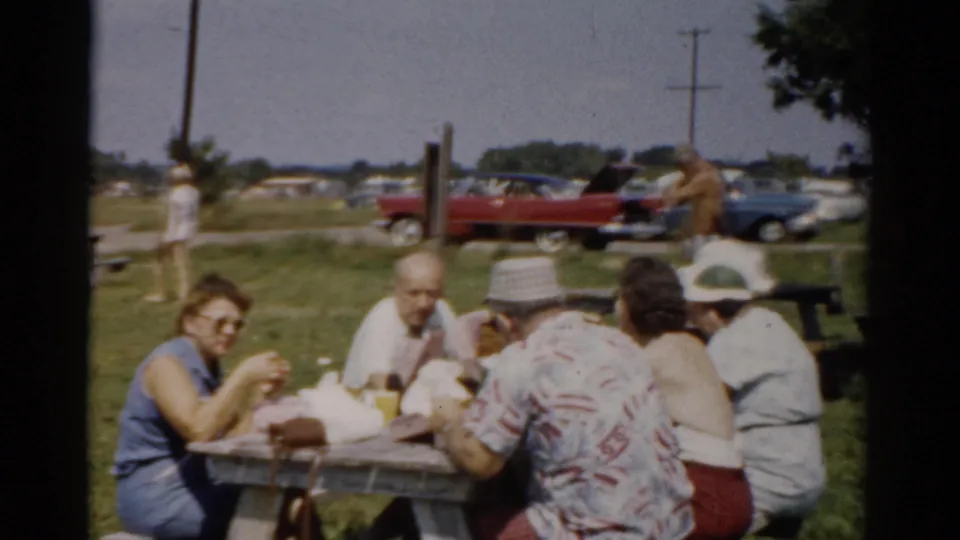 Adults sit around a wooden picnic table at a Catskill, New York outdoor gathering, with food wrappers, hats, and 1950s cars visible behind them.