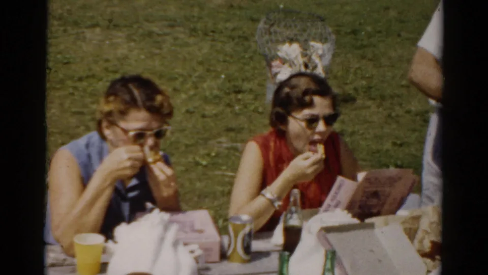 Close home-movie view of two women eating at a picnic table in 1957 Catskill, with sunglasses, bottles, paper cups, and a printed program visible.
