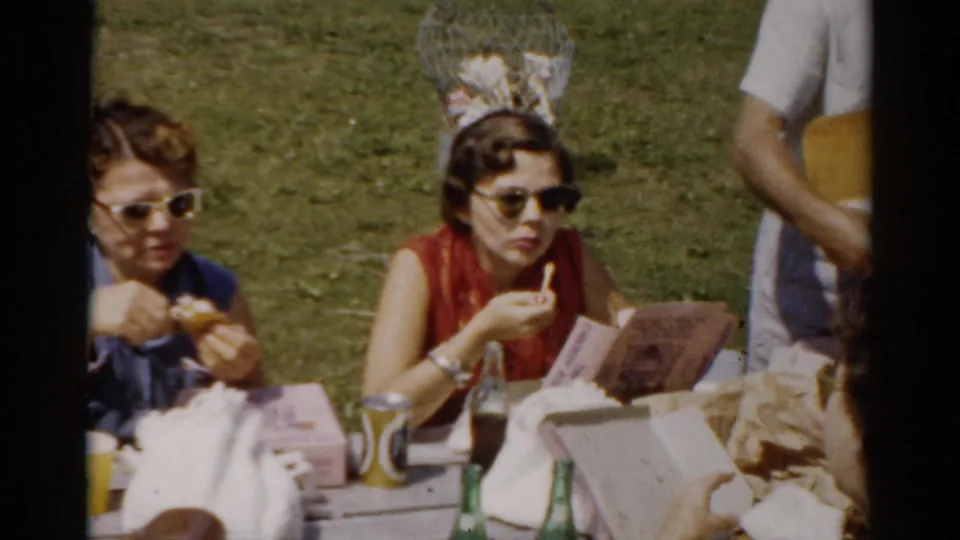 Catskill, New York picnic close-up showing women in sunglasses eating at a wooden table with bottles, cups, food bags, and a program in bright daylight.