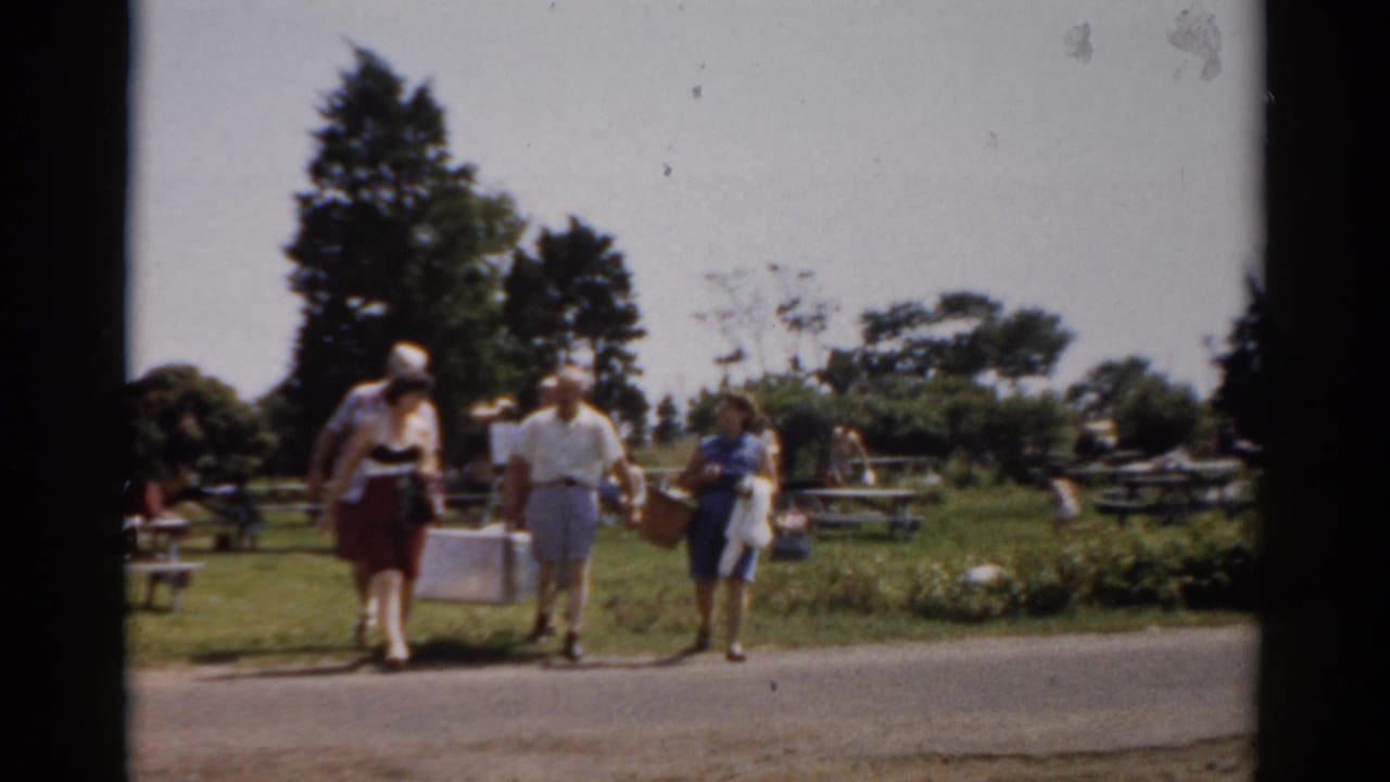 In Catskill, New York home-movie footage from 1957, adults in summer clothing walk across a road with a cooler and basket near park picnic tables.