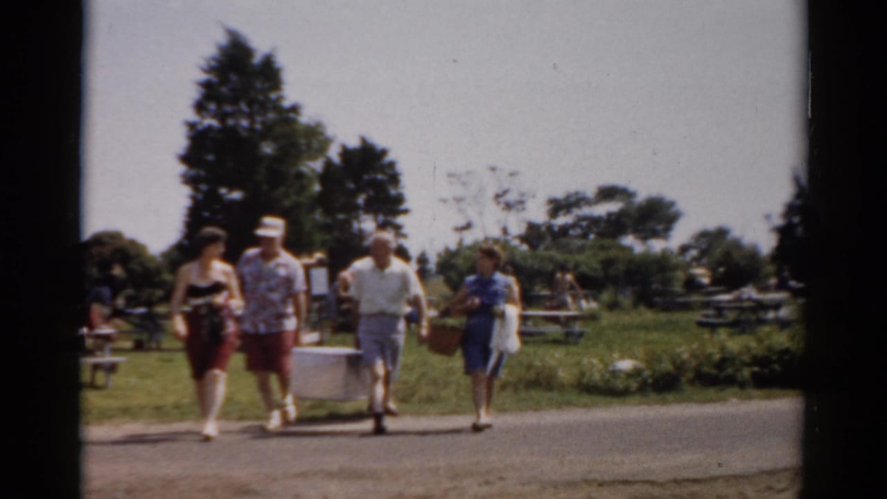 Adults in 1950s summer attire cross a road in Catskill, New York, carrying a white cooler and picnic basket through a park setting.