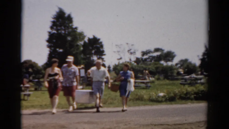 Adults in 1950s summer attire cross a road in Catskill, New York, carrying a white cooler and picnic basket through a park setting.