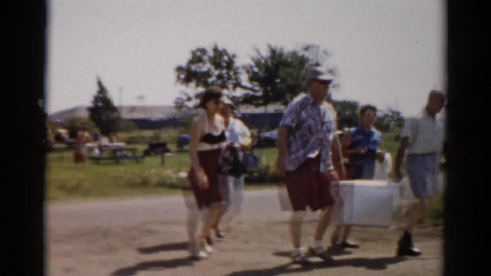 The family group passes nearer the camera, with a man carrying a white cooler and others holding bags and a picnic basket in a grassy park area.