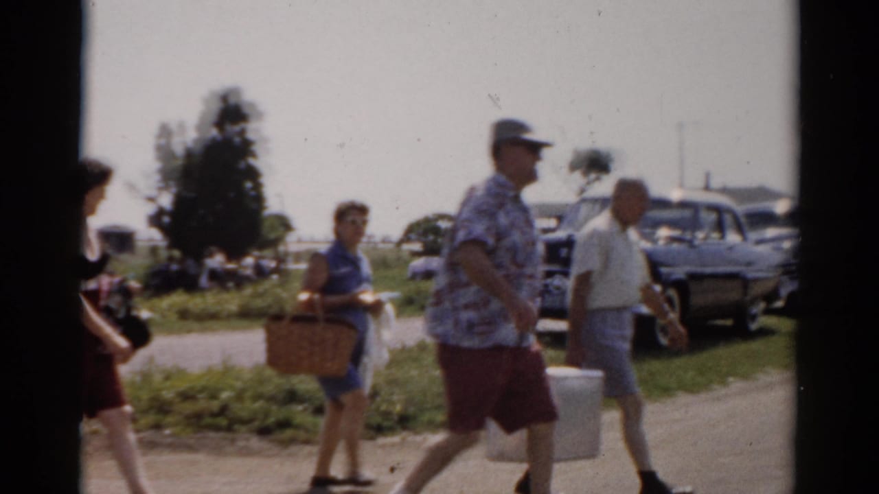 In Catskill, New York, adults in summer clothing carry picnic baskets and a cooler past parked mid-century cars in 1957 home-movie footage.