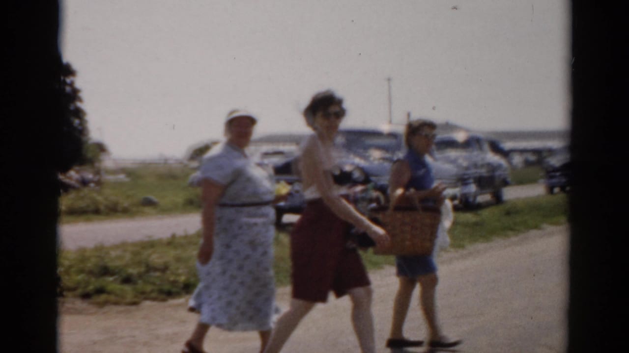Adults in summer outfits walk rightward with woven picnic baskets along a Catskill roadside, with 1950s cars parked behind them.