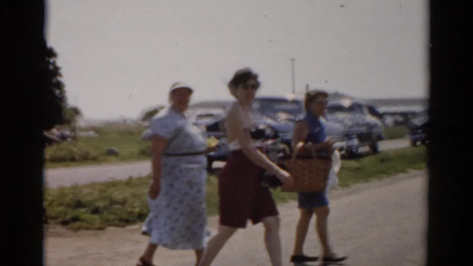 Adults in summer outfits walk rightward with woven picnic baskets along a Catskill roadside, with 1950s cars parked behind them.