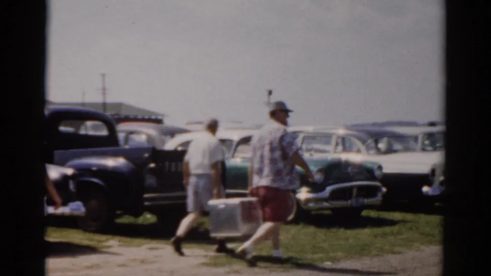 Two men move across a grassy roadside with a white cooler, with mid-century cars parked behind them in the supplied 1957 Catskill setting.